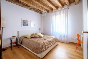 A bedroom with a wooden ceiling, a bed with a beige duvet, a bedside table with books, a lamp, and a window with curtains.