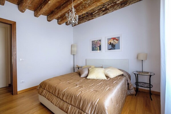 A bedroom with a wooden ceiling, a bed with a beige comforter, two wall-mounted lamps, and two framed pictures on the wall.