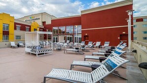 Un espace de détente sur le toit, avec des chaises longues et un coin salon couvert, un bâtiment à la façade rouge et une enseigne indiquant « GreenVacations ».