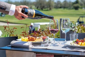 A person pouring wine into a glass on an outdoor table with plates of food, glasses, and a bottle of wine.