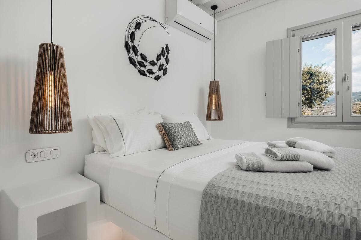 A modern bedroom with a bed, bedside tables, a wall-mounted air conditioner, and a window with a view.