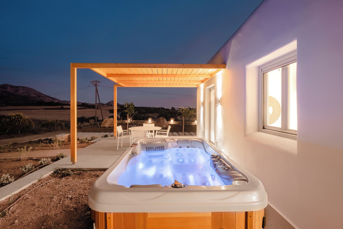 A hot tub with a wooden pergola, a patio table, and chairs, set against a white wall with a window.