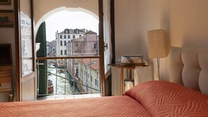 Ein Hotelzimmer mit Bett, kleinem Tisch mit Lampe und Blick auf einen Kanal sowie Gebäude durch eine geöffnete Tür.