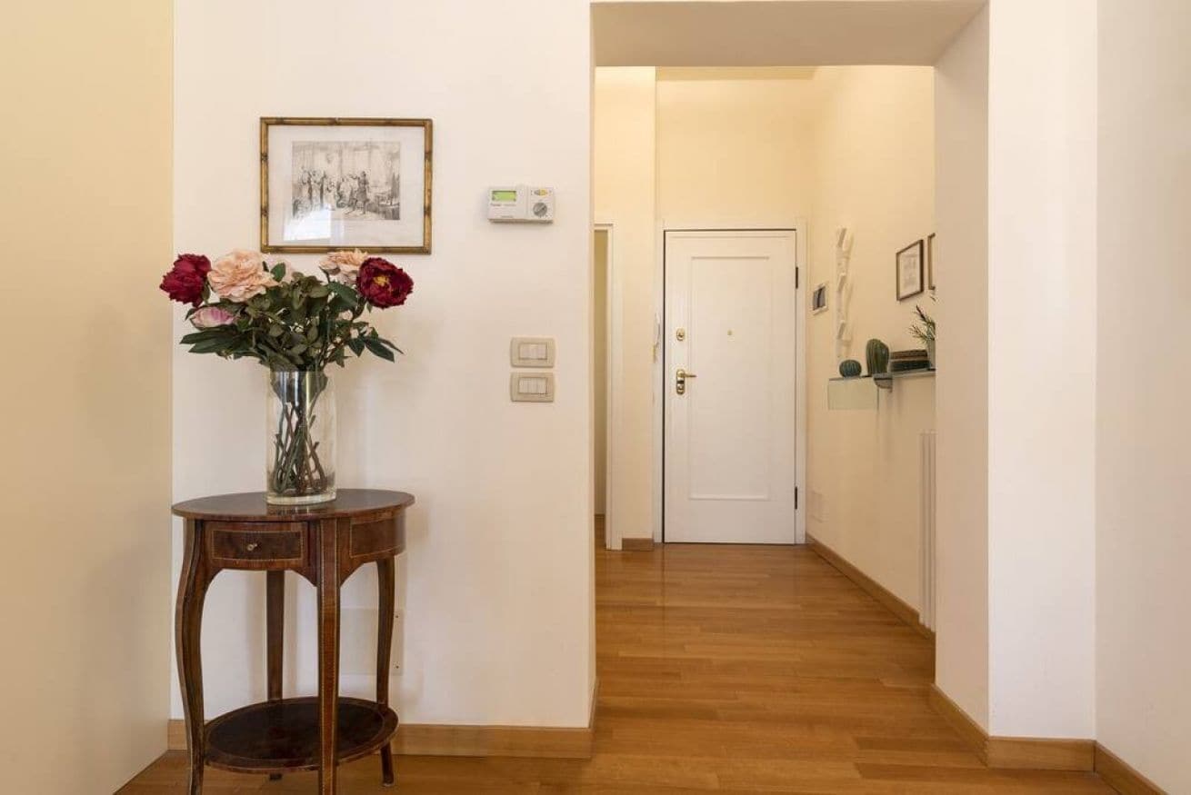 Un corridoio con pavimento in legno, una porta bianca in fondo, un tavolino con un vaso di fiori e un quadro appeso al muro.