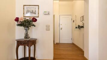 Un corridoio con pavimento in legno, una porta bianca in fondo, un tavolino con un vaso di fiori e un quadro appeso al muro.