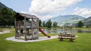 A rustic wooden playhouse with a slide, situated on a grassy area near a lake with mountains in the background. - Mountain-view Cottage 15 Beds-pets Welcome (Bramberg am Wildkogel)