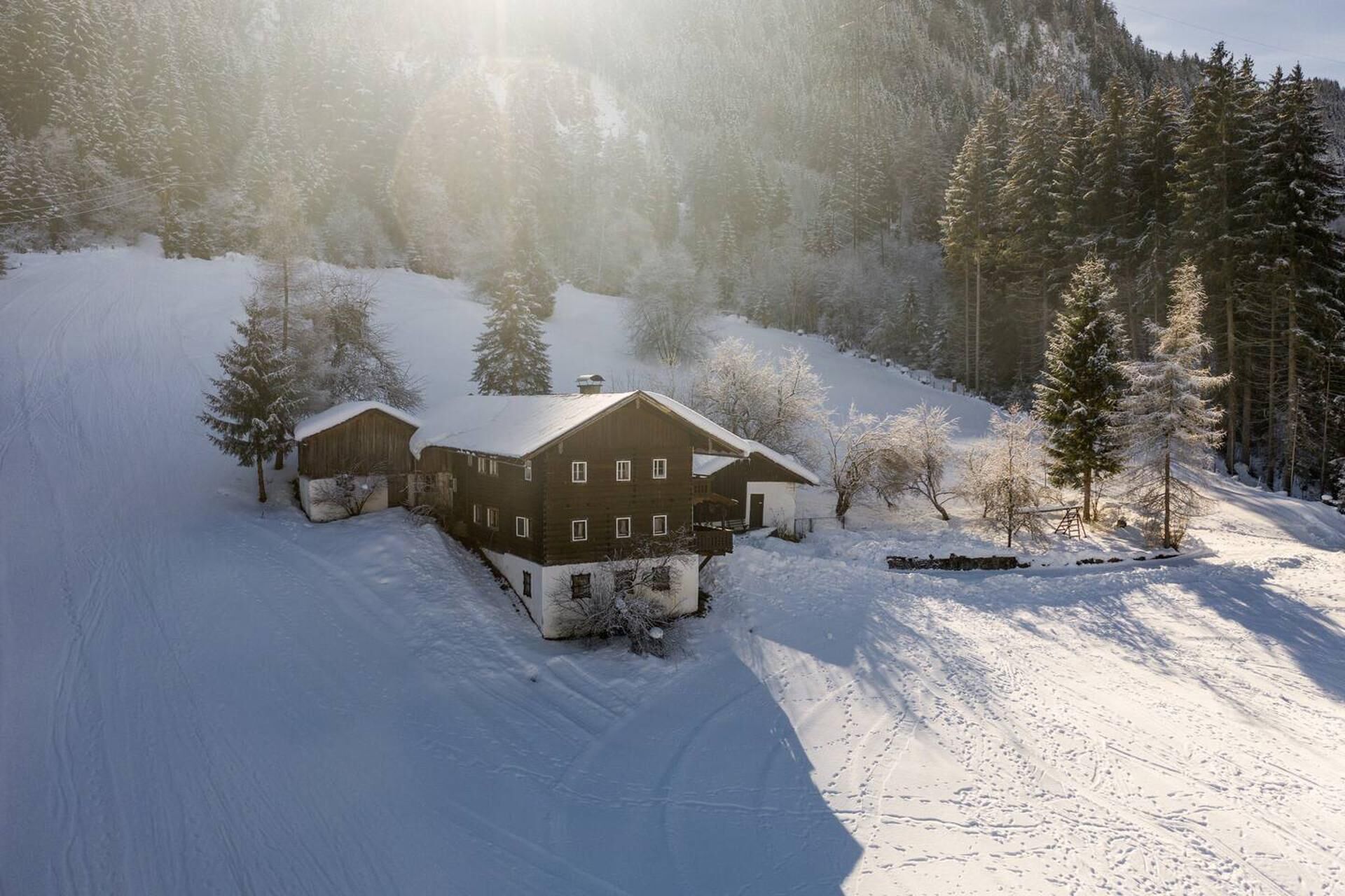 A wooden cabin with a sloped roof, surrounded by snow-covered trees and a mountain backdrop.