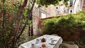 A table set for two with a floral tablecloth, a teapot, cups, and a plate of cookies in a courtyard with brick walls and greenery.