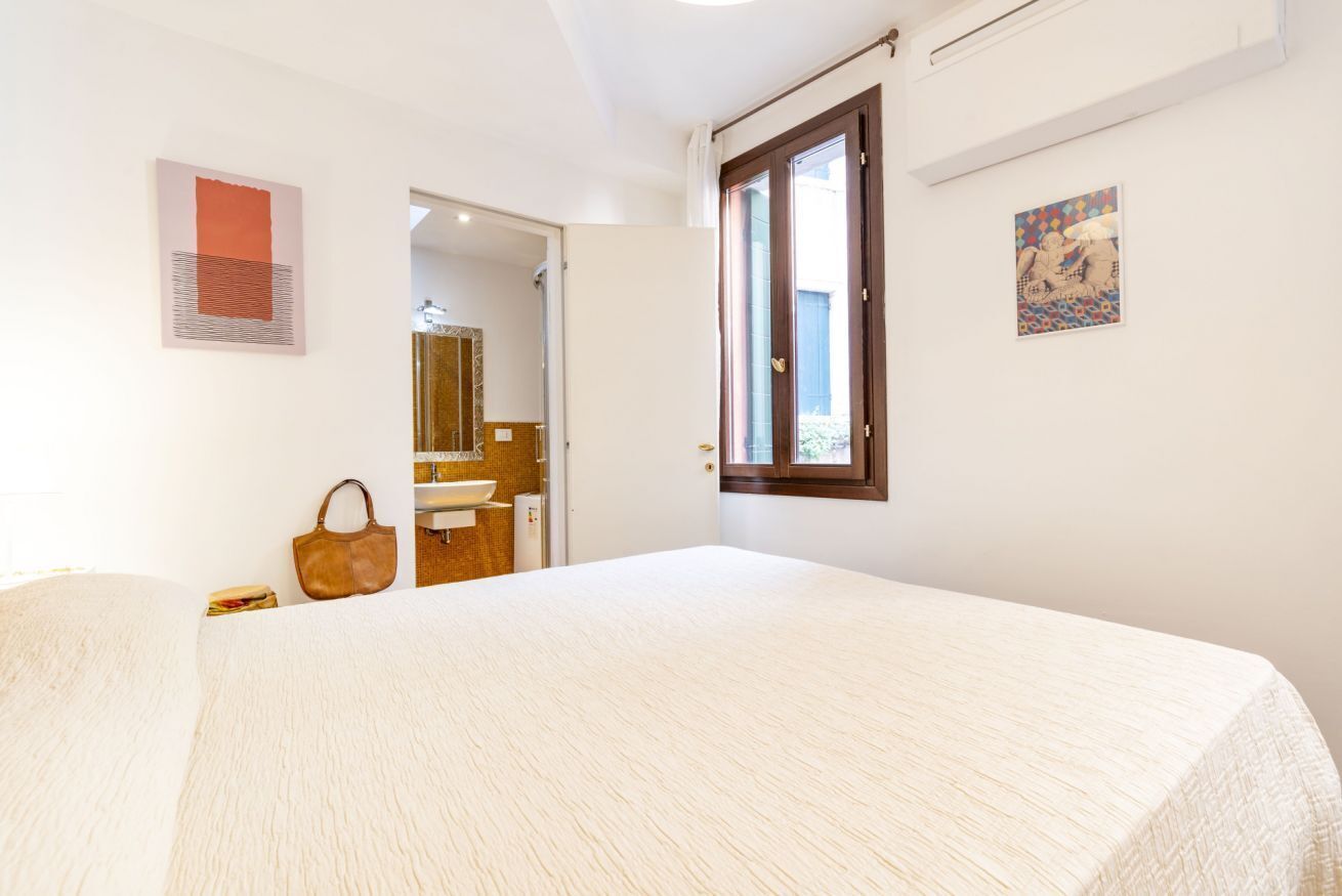 Camera da letto con un letto grande, una porta in legno, una finestra e un bagno visibile attraverso una porta aperta.