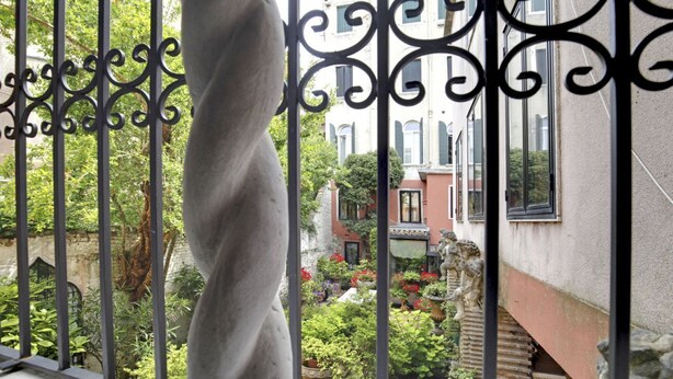 Blick durch kunstvolle Metallgitter auf einen Innenhof mit Statue, Garten und Gebäuden im Hintergrund.