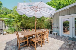 An outdoor patio with a wooden table and chairs, a striped umbrella, and a view of a garden and house.
