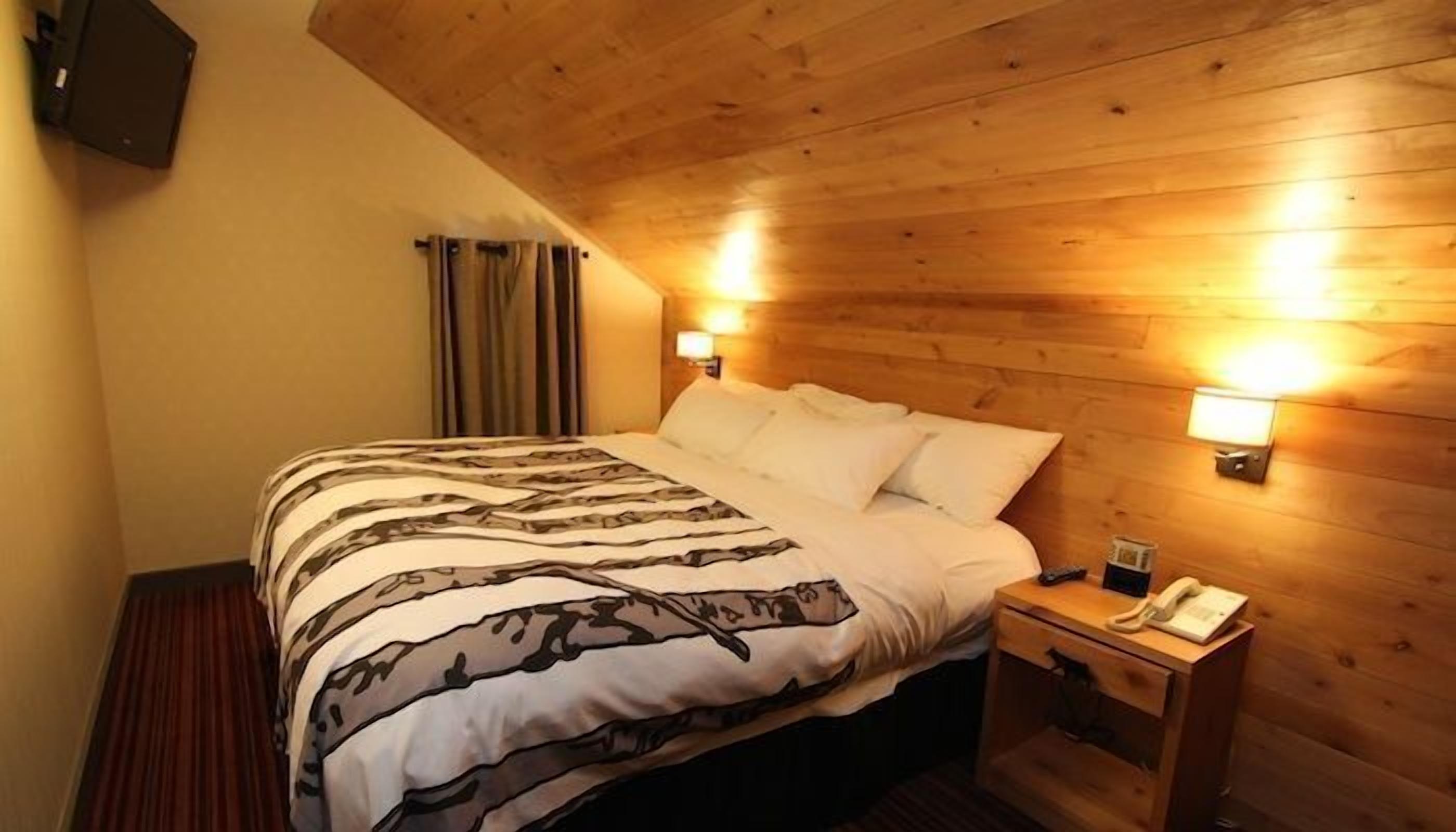 A hotel room with a wooden ceiling, a bed with patterned bedding, two bedside lamps, and a nightstand with a phone.