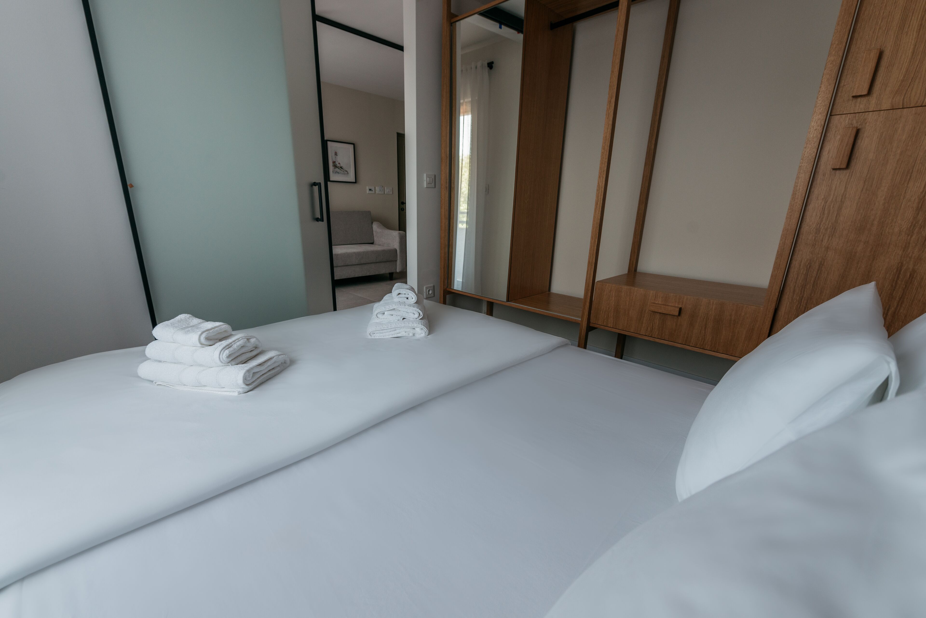 A neatly made bed with white linens and towels, a wooden wardrobe, and a mirror.