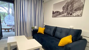 A modern living room with a dark blue sofa, two yellow cushions, a white coffee table, and a black rug. There is a large window with blue and white striped curtains, a hanging chair, and a black and white landscape painting on the wall. - Family Resort Urania (Baska Voda)