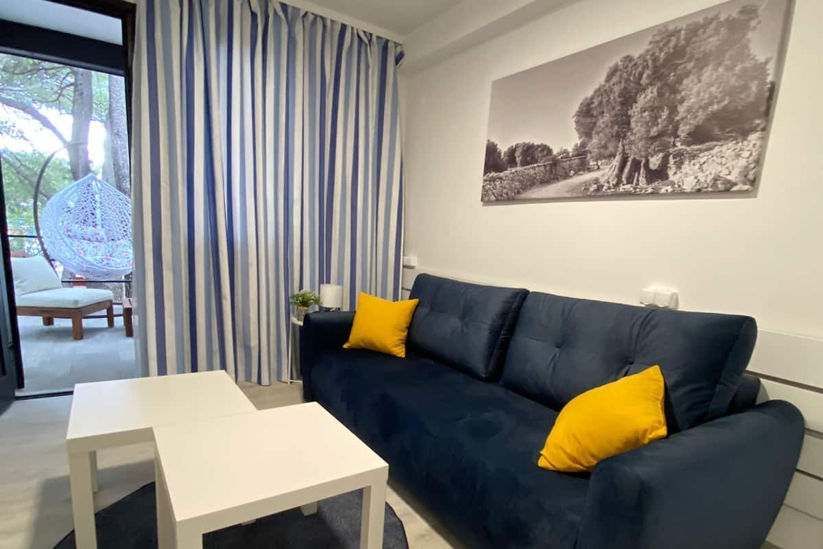 Uma sala de estar moderna com um sofá azul escuro, duas almofadas amarelas, uma mesa de centro branca e um tapete preto. Há uma janela grande com cortinas listradas em azul e branco, uma cadeira suspensa e uma pintura de paisagem em preto e branco na parede.
