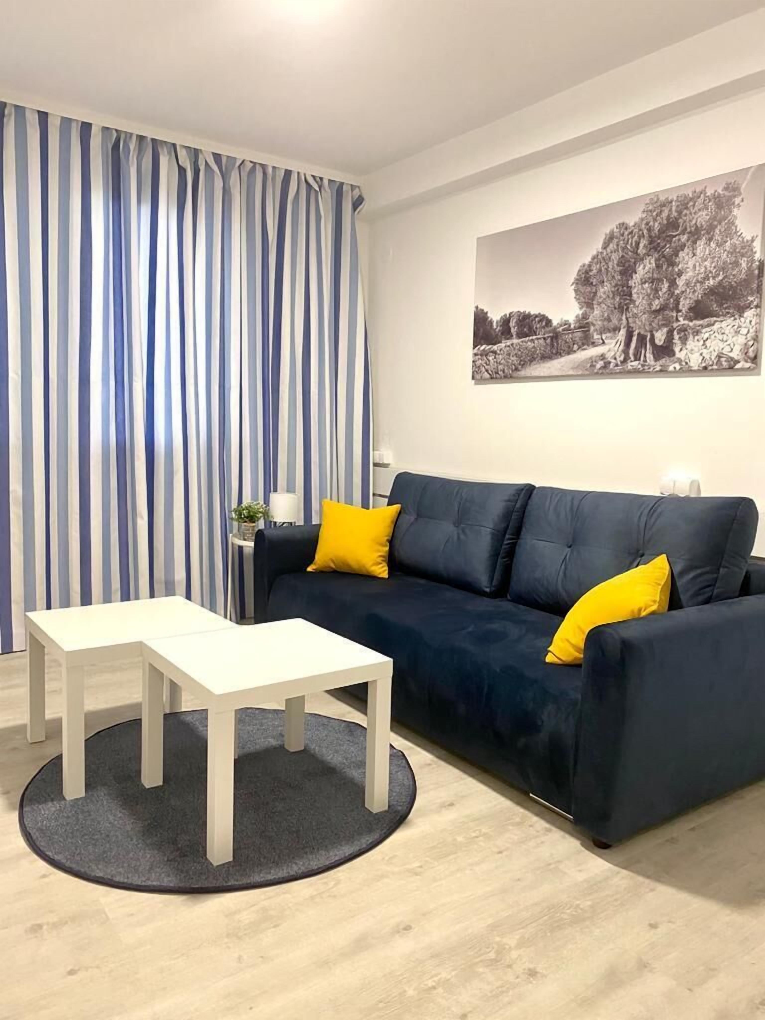 Un salon moderne avec un canapé bleu foncé, deux coussins jaunes, une table basse blanche et un tapis gris. Un rideau à rayures et une photographie en noir et blanc ornent les murs.