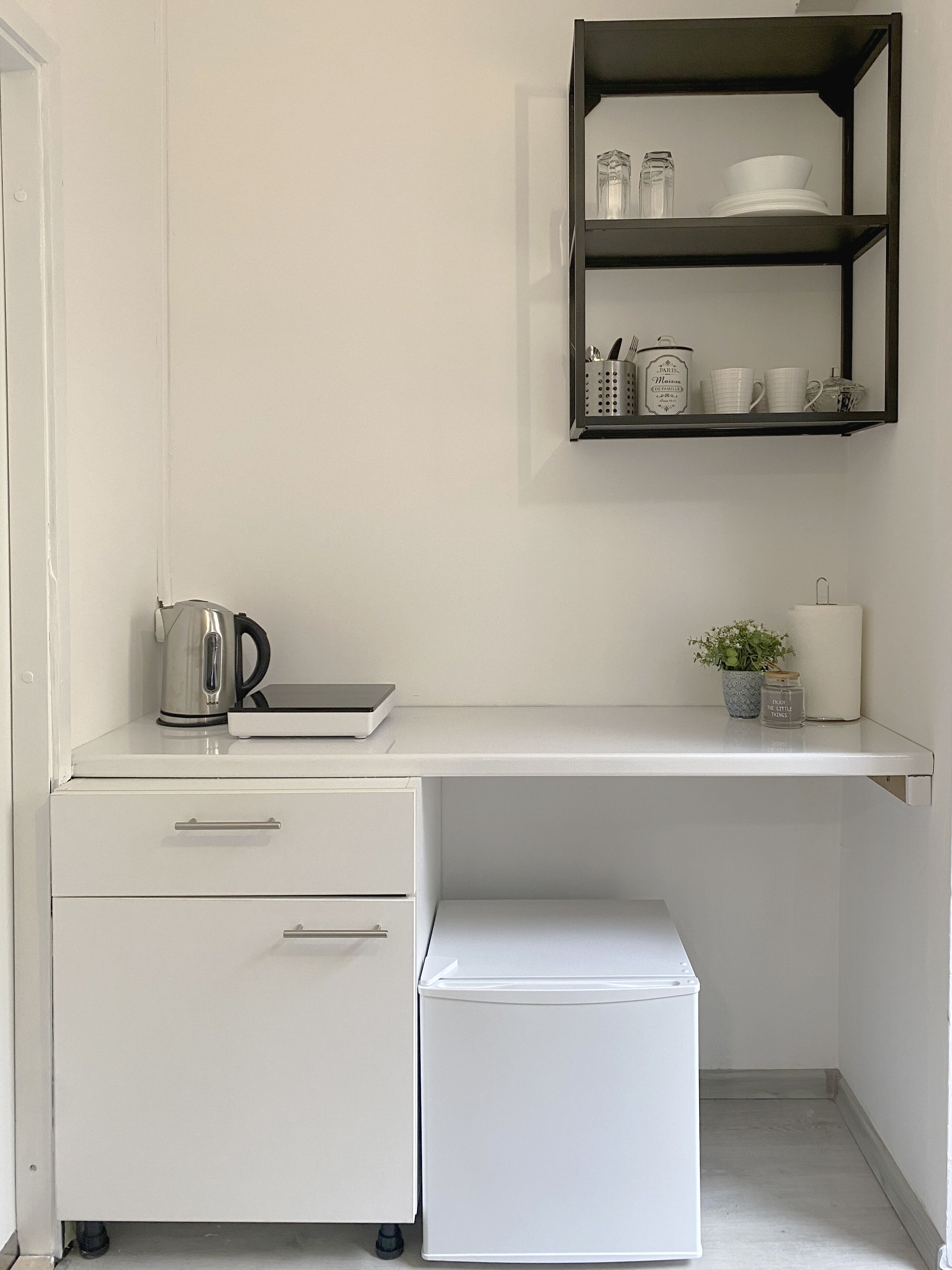 A kitchenette with a white countertop, a kettle, a toaster, a small refrigerator, and a shelf with kitchenware.