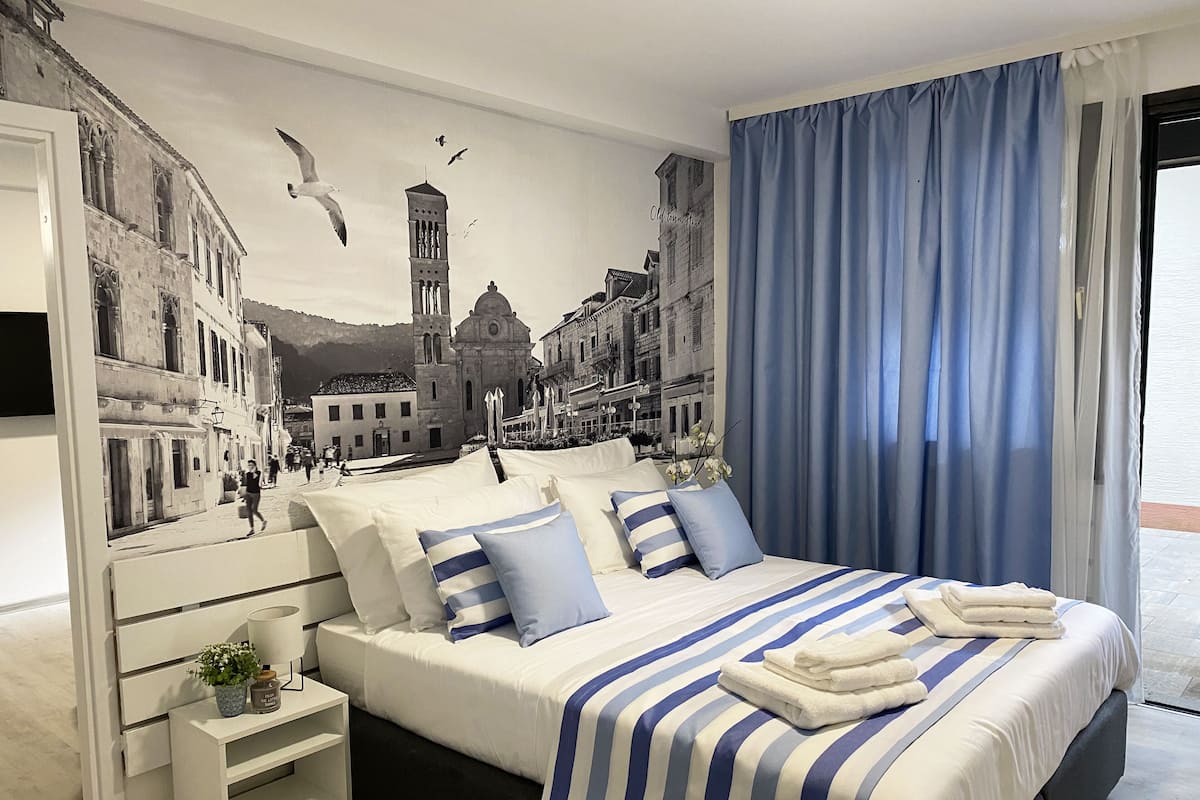 Quarto com cama, almofadas, mesa de cabeceira, abajur e um mural na parede com vista urbana.