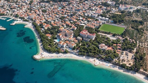 An aerial view of a coastal town with a beach, residential buildings, and a sports field. - Family Resort Urania (Baska Voda)