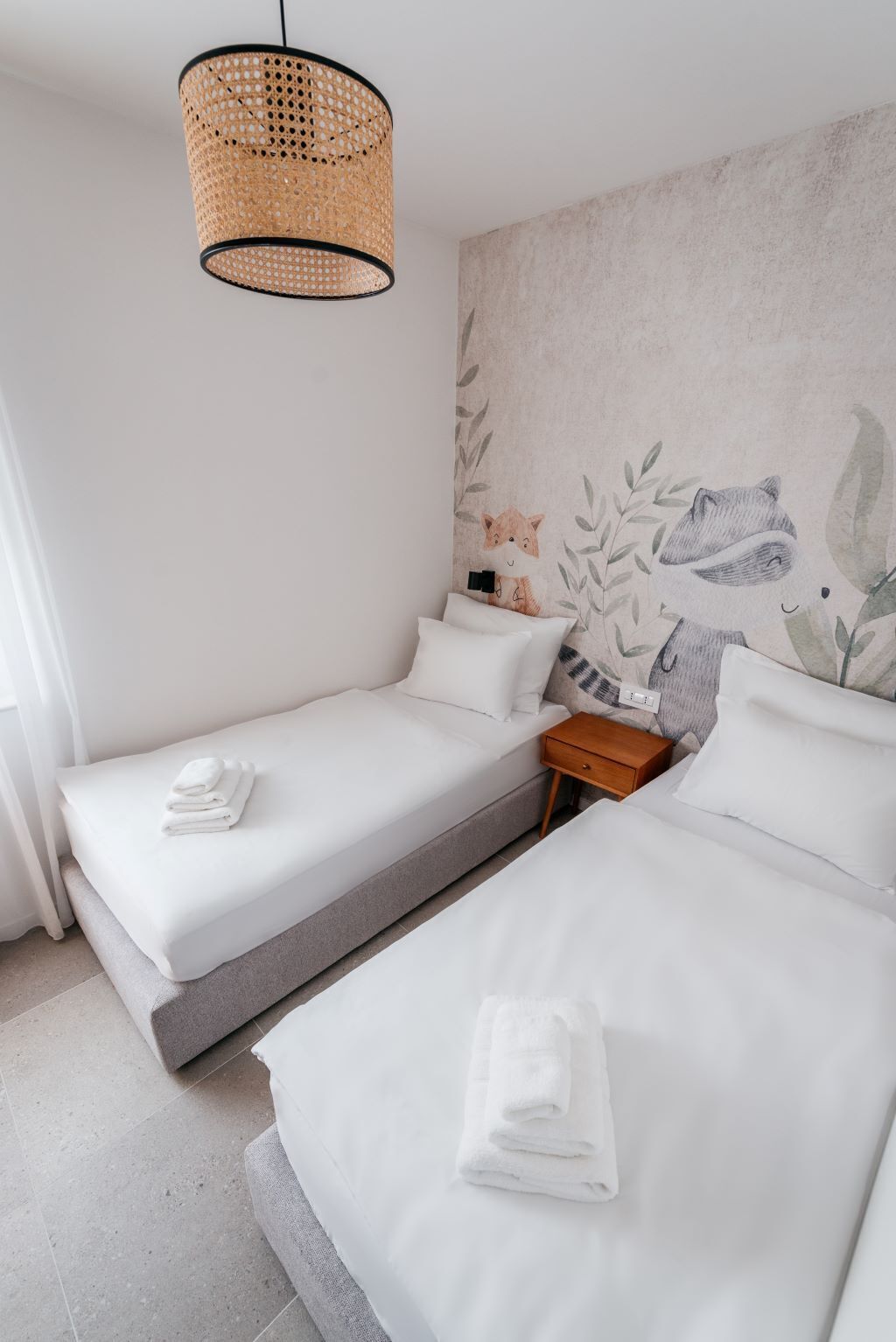 A modern hotel room with two single beds, a woven pendant light, a nightstand, and a wall mural.