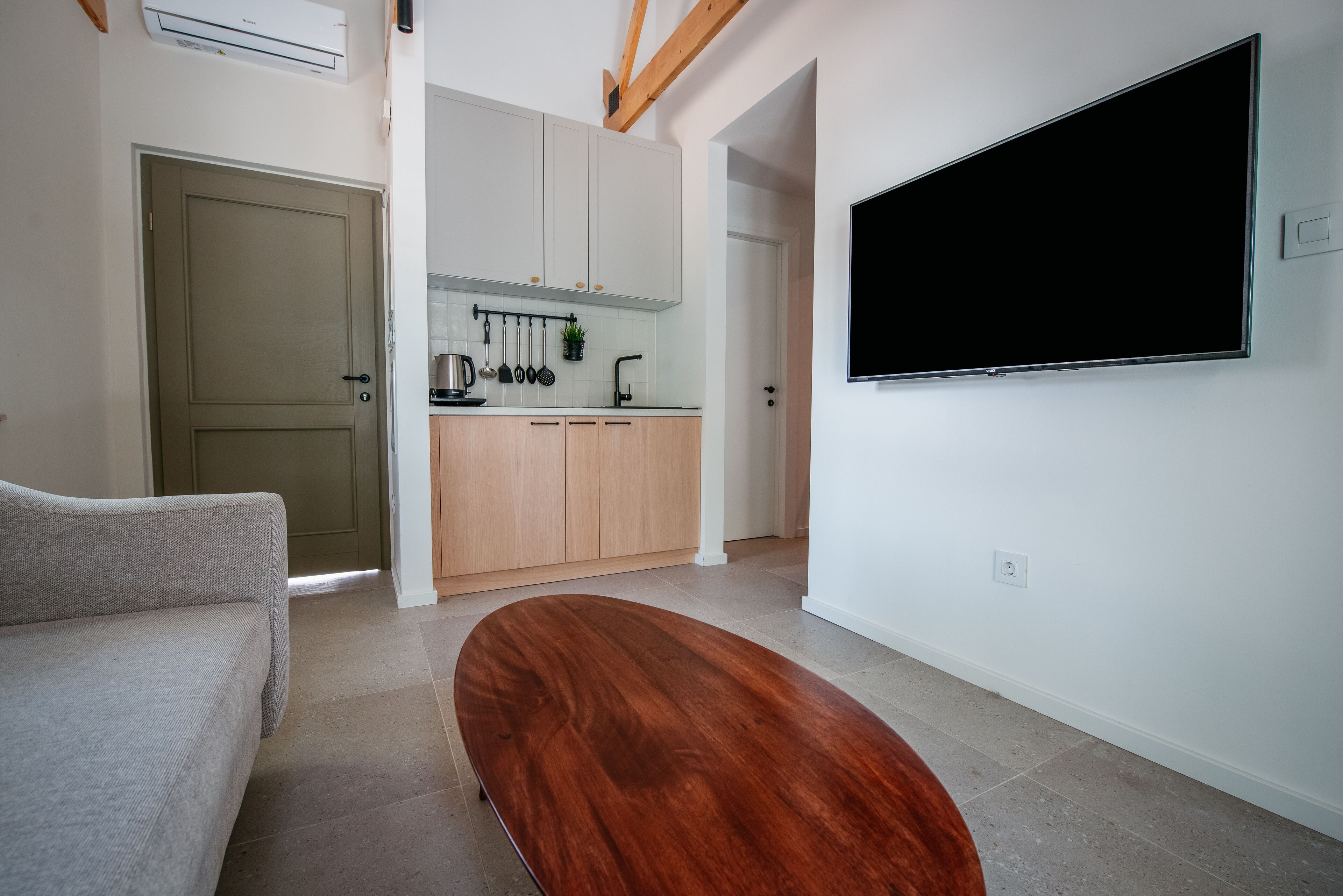 A modern living room with a wooden coffee table, a grey sofa, and a kitchen area with white cabinets and a flat-screen TV mounted on the wall.