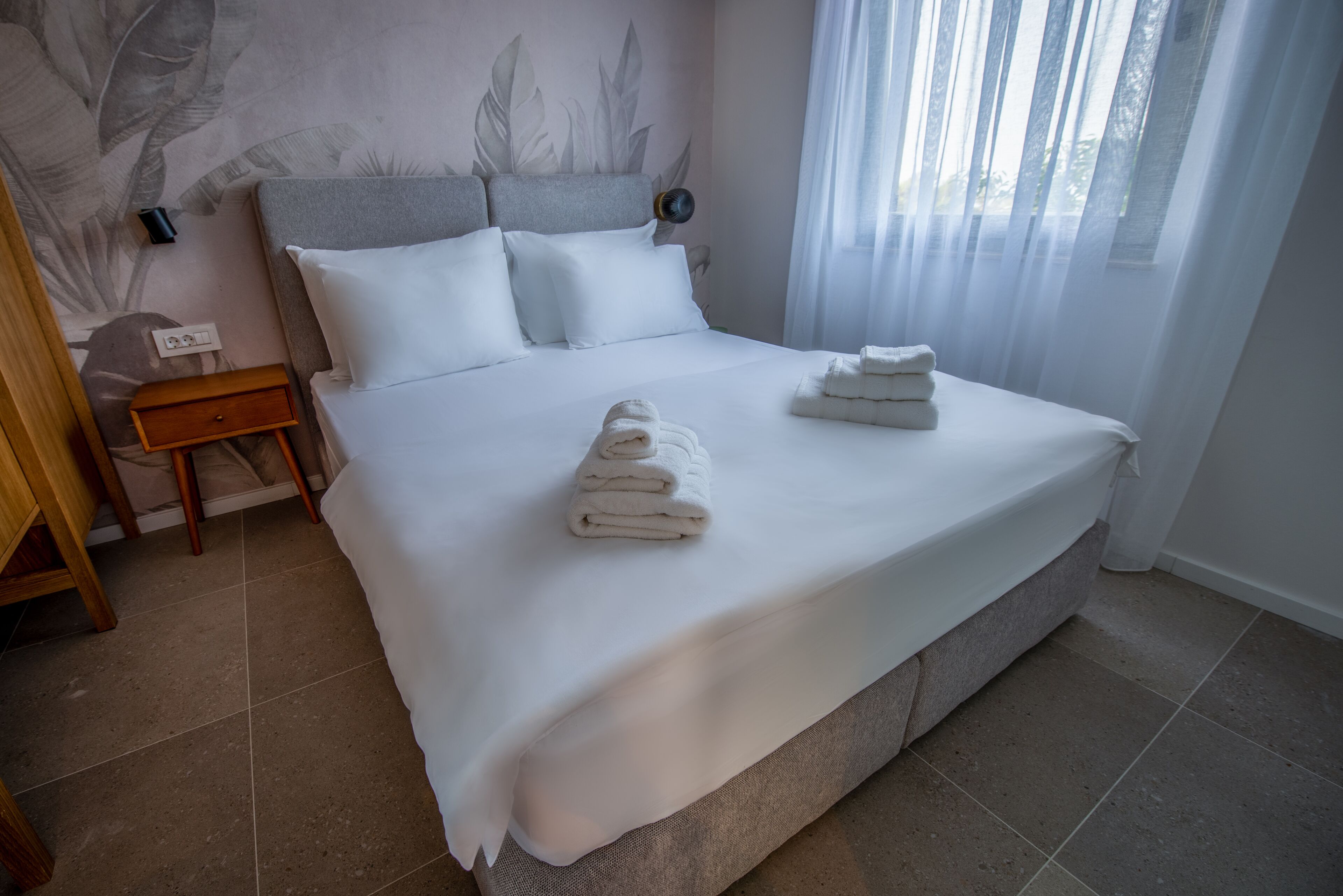A neatly made bed with white linens and towels, a bedside table with a lamp, and a wall with a leaf pattern.