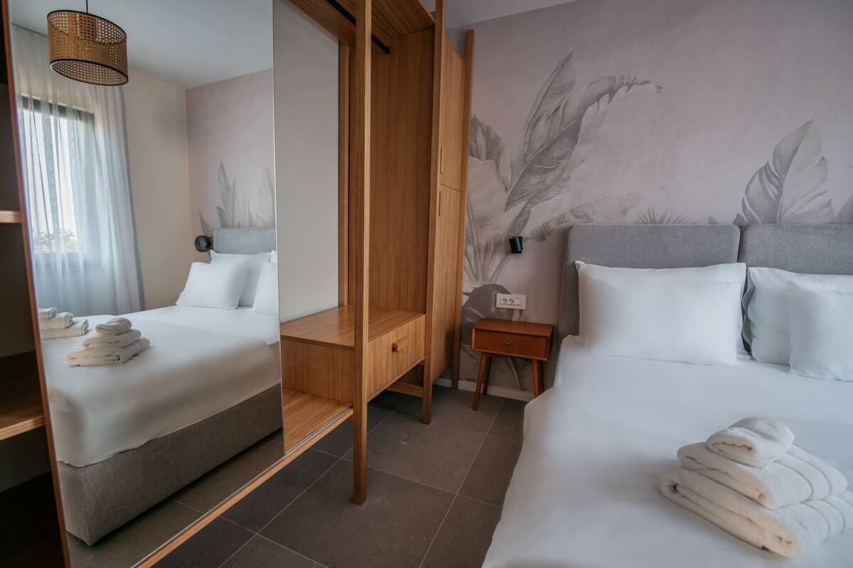 Quarto de hotel com duas camas, um guarda-roupa de madeira, uma mesa de cabeceira pequena e uma parede com padrão de folhagens.