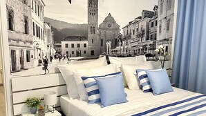 A bedroom with a mural of a cityscape, a bed with striped bedding, a nightstand with a lamp, and a view of the sky. - Family Resort Urania (Baska Voda)
