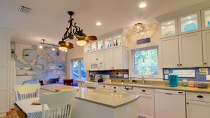 A kitchen with white cabinets, a map on the wall, a dining area with a table and chairs, and a window with a view of trees. - Whimsea: West End, Pets OK, Pool Home on Canal! (Sanibel)