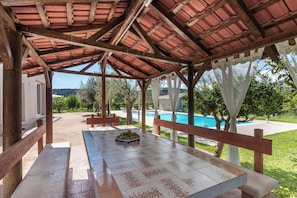 An outdoor covered patio with a pool, wooden benches, and a tiled table.