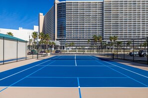 Un court de tennis en plein air, situé à côté d’un hôtel, avec un immeuble de grande hauteur en arrière-plan.