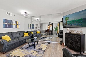 Una sala de estar moderna con un sofá de cuero negro, una mesa de centro y un espejo grande que refleja la habitación. Hay un cuadro en la pared y un cartel de Emerald Beach Properties.