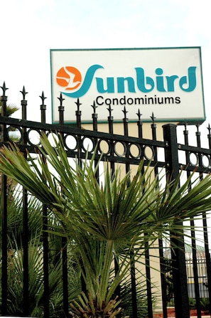 Schild für die Sunbird Condominiums mit dekorativem Zaun und Palmen im Vordergrund.