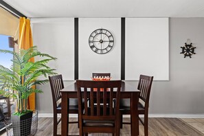 A dining area with a wooden table and chairs, a wall-mounted clock, and a potted plant. - Seaside Serenity Sunbird 503W-Gorgeous Beach Views! Comp Beach Service! (Panama City Beach)