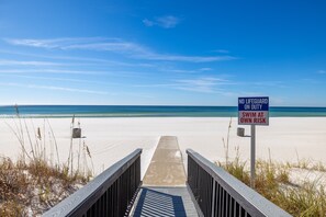 A beach with a wooden boardwalk leading to the water, a lifeguard sign, and clear skies.