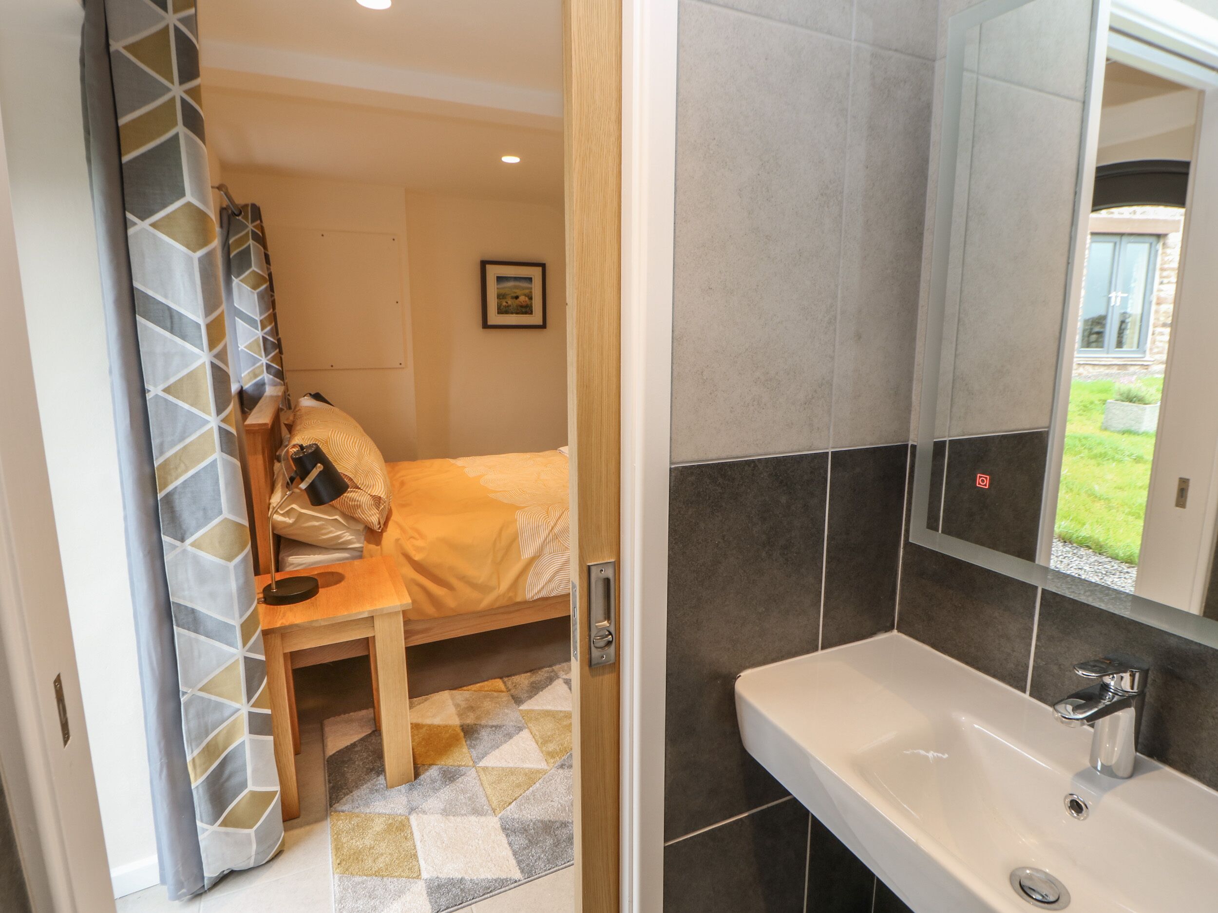 Ein Hotelzimmer mit Bett, Nachttisch aus Holz, Bad mit Waschbecken und Spiegel sowie Blick ins Freie.