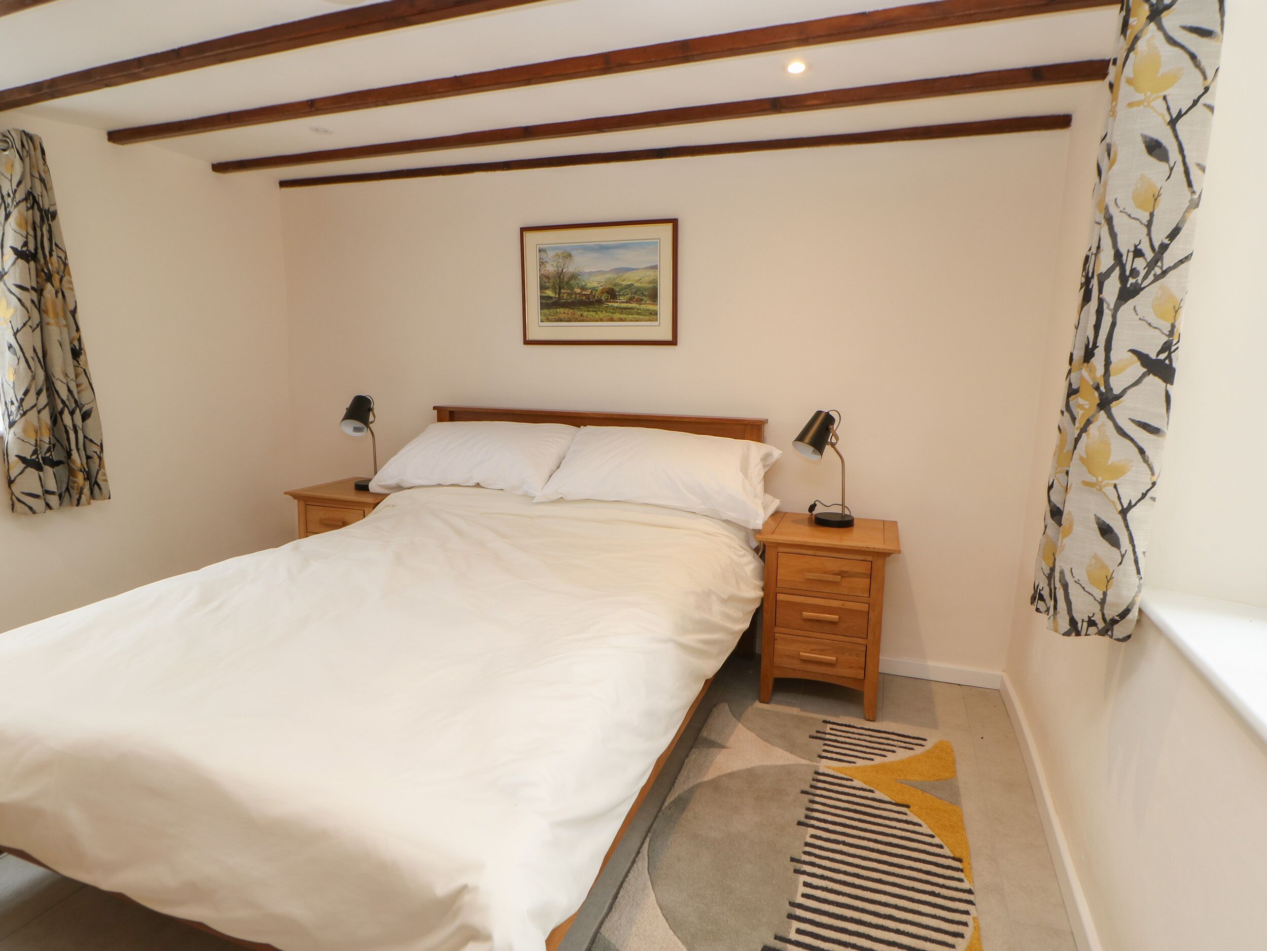 A bedroom with a large bed, wooden headboard, two bedside tables with lamps, a framed picture on the wall, and patterned curtains.