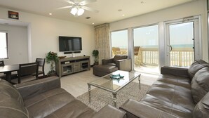 Un salon spacieux avec un grand canapé, une table basse et un téléviseur. Il y a un balcon avec vue sur la plage.