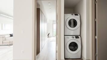 A laundry room with two stacked washing machines, a hallway leading to a living area with a sofa, and a door to another room.