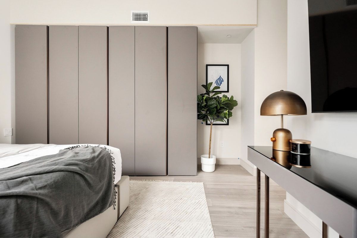 A bedroom with a bed, a wardrobe, a desk with a lamp, and a potted plant.