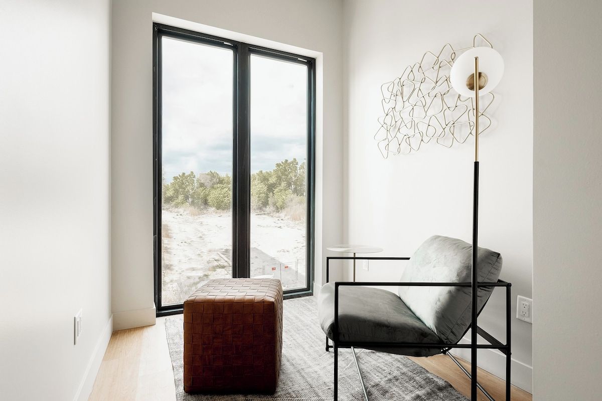 A modern room with a large window, a floor lamp, a grey chair, and a textured ottoman.