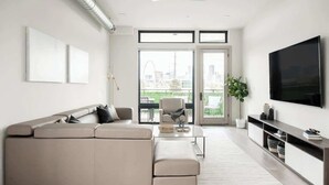 A modern living room with a beige sectional sofa, a flat-screen TV, and a large window offering a city view. - Denver Skyline Views | Walk to Empower Field (Denver)