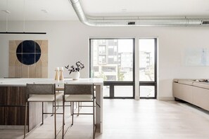 A modern kitchen with a central island, bar stools, and a view through a glass door to an outdoor balcony. - Denver Skyline Views | Walk to Empower Field (Denver)