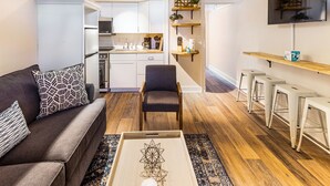 Un espai habitable compacte amb cuina americana, un sofà amb coixins estampats, una taula de centre amb disseny geomètric i un televisor penjat a la paret.