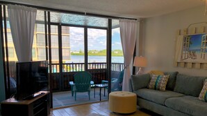 A living room with a television, a sofa, a chair, and a table. There is a balcony with a view of the outside. - 4 Minute Walk to Beach! Waterfront and Pool View-Indian Shores Beach! (Indian Shores)