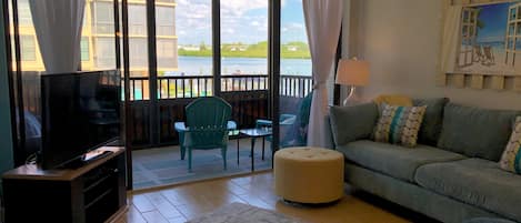A living room with a television, a sofa, a chair, and a table. There is a balcony with a view of the outside.