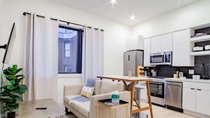Una sala de estar moderna con un sofá, una mesa de centro y una mesita auxiliar. Se observa una zona de cocina con microondas, horno y lavavajillas. Una ventana grande con cortinas es visible.