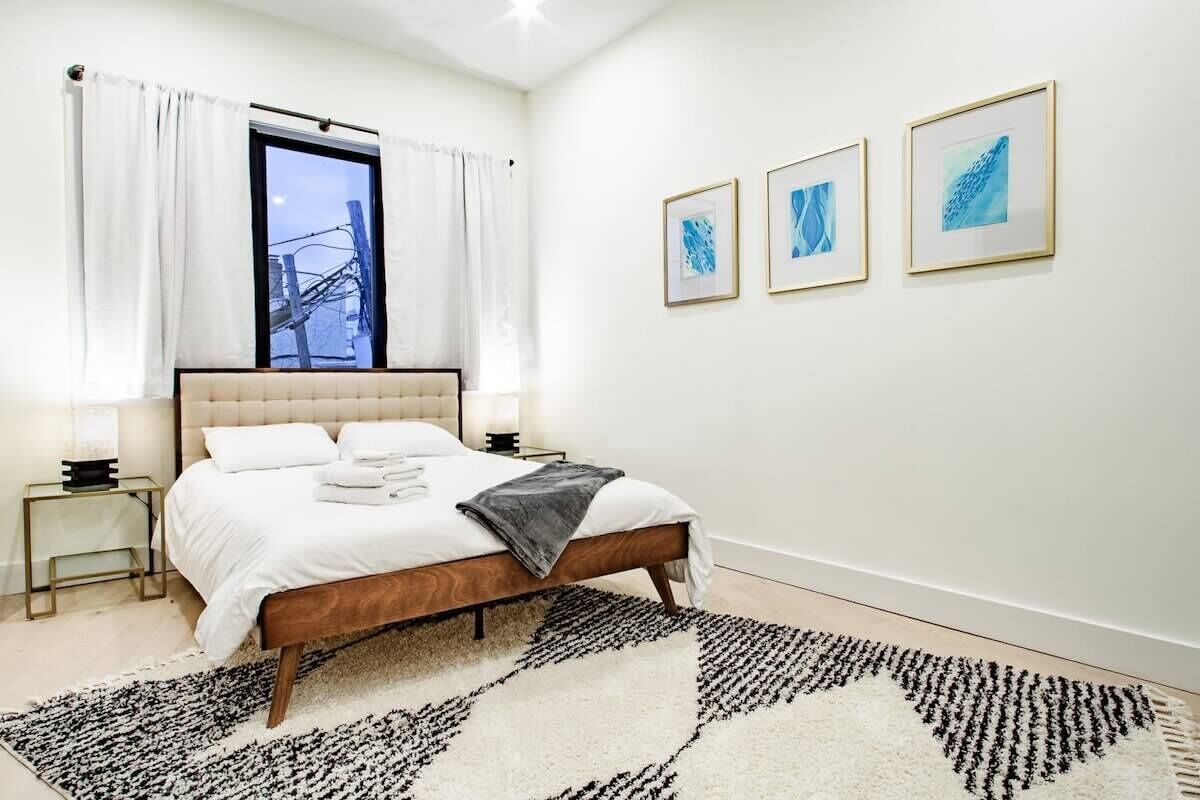 Un dormitorio con cama, mesitas de noche, una ventana con cortinas y una obra de arte en la pared.