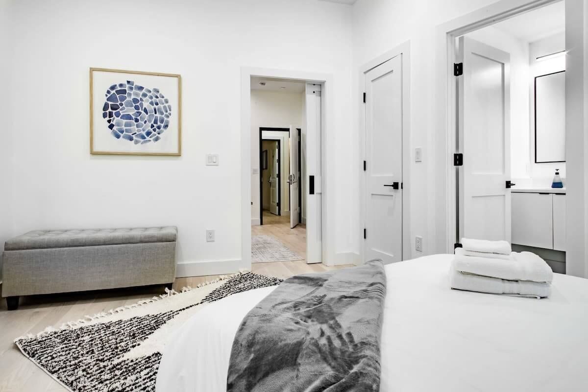 A modern bedroom with a bed, a grey bench, a rug, and a framed artwork on the wall.