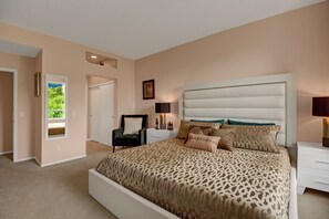 A bedroom with a large bed, a chair, a nightstand, and a window with a view of greenery. - Fully Renovated and Furnished Regency Style Condo in La Palme (Palm Springs)